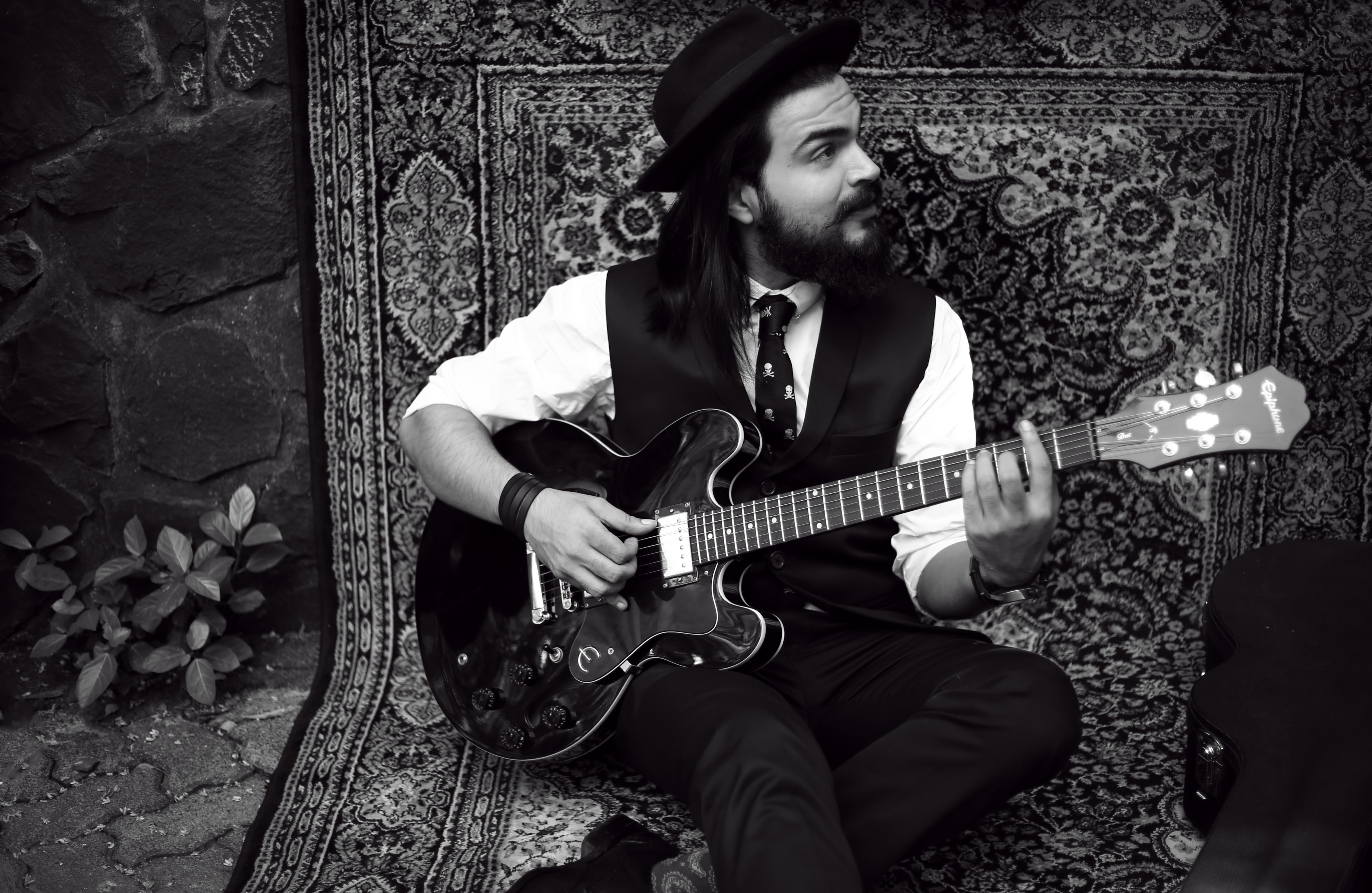 Mumbai-based singer, songwriter, and guitarist Umang Mehta playing an Epiphone electric guitar while seated on a patterned rug, wearing a hat, vest, and skull-print tie in a vintage, artistic outdoor setting.