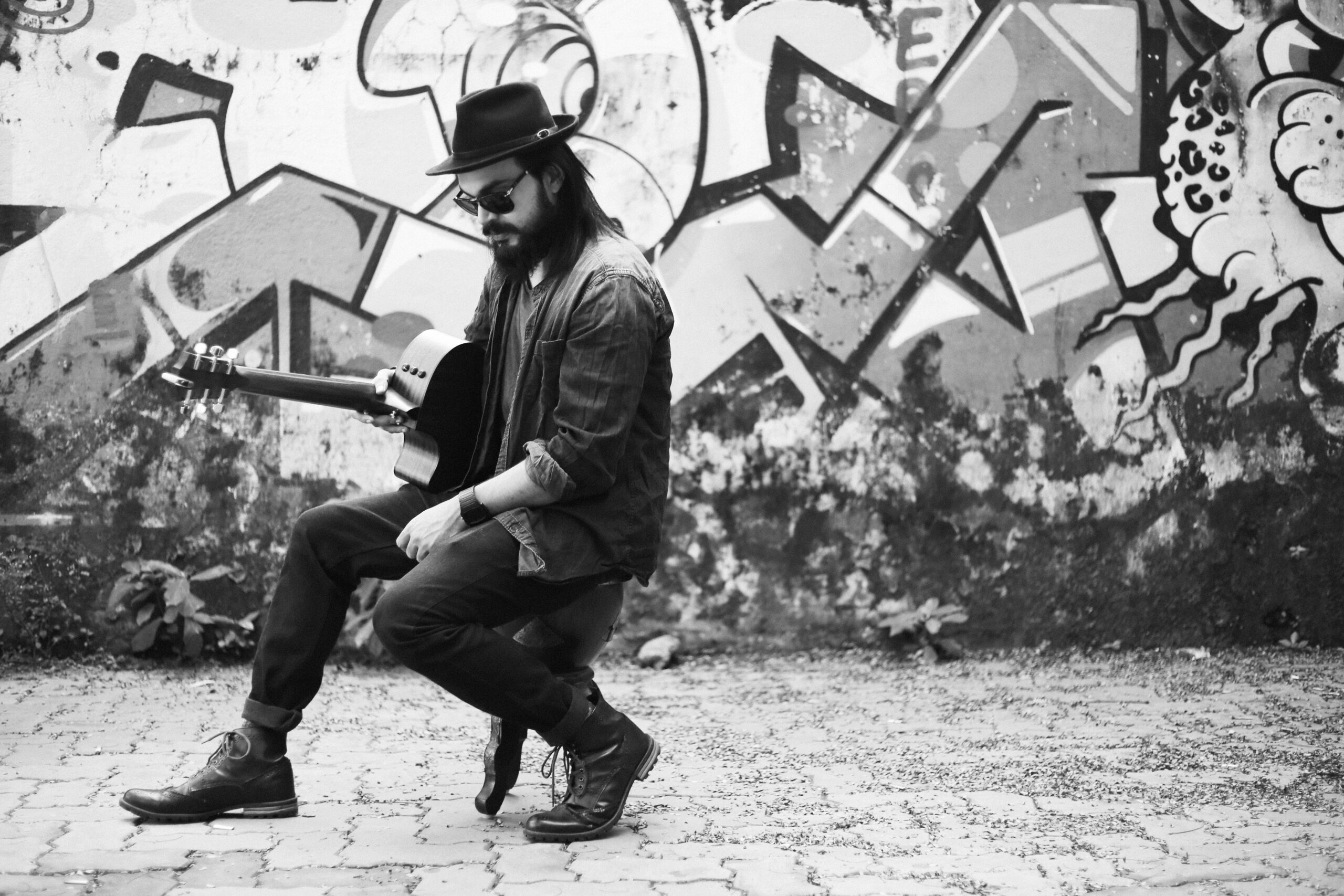 Mumbai-based singer, songwriter, and guitarist Umang Mehta sitting with an acoustic guitar in front of a colorful graffiti wall, wearing a fedora, sunglasses, and boots in a relaxed urban street-style setting.